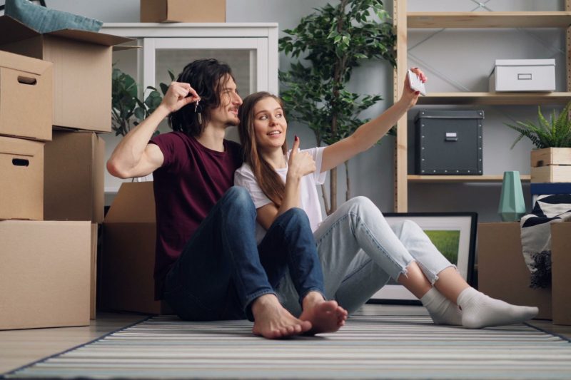 Young couple sitting among moving boxes, taking a selfie, celebrating new home